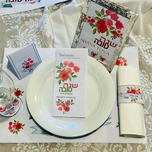 May include: A festive table setting with a white plate, napkin, and glass. Floral accents in red and pink adorn the place setting, including a coaster, place card, and napkin ring. A framed card reads "Shana Tova."