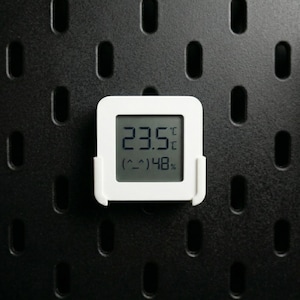 Puede incluir: Un termómetro digital blanco que muestra 23,5°C y 48% de humedad, montado en una placa perforada negra. El termómetro tiene una pantalla cuadrada con esquinas redondeadas y un pequeño soporte.