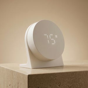 May include: A white, modern-style digital clock displaying "75°" on a circular screen. The clock has a minimalist design with a rounded face and a rectangular base. The clock is sitting on a stone surface.