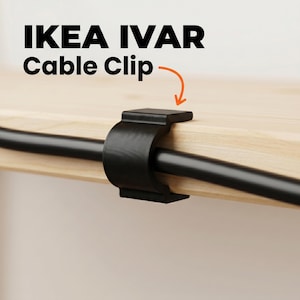 Puede incluir: Un clip para cables de plástico negro sujeto a un estante de madera clara, asegurando un cable negro. El texto IKEA IVAR Cable Clip está impreso encima con una flecha naranja que apunta hacia el accesorio de gestión de cables negro.