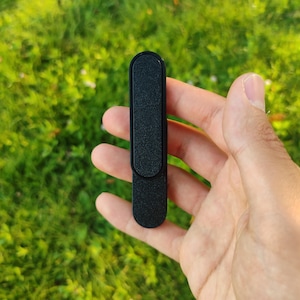 Op de afbeelding: Een zwarte, rechthoekige telefoongreep met een textuuroppervlak. De grip wordt tegen een achtergrond van groen gras gehouden.