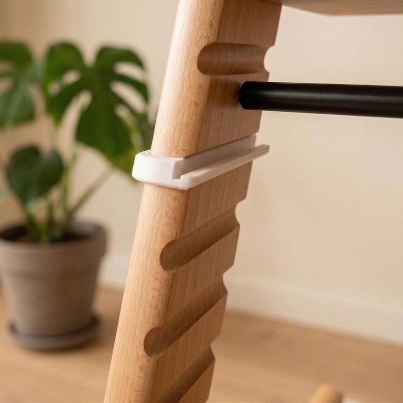 May include: Close-up of a wooden high chair leg with a black bar and white plastic support. The chair leg has a natural wood grain finish and a series of curved cutouts. A potted plant is visible in the background.
