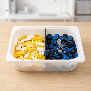 Könnte beinhalten: Eine weiße, geteilte Kunststoffbox gefüllt mit gelben, weißen und blauen Spielzeugbausteinen und Rädern. Die Box ist in zwei Abschnitte unterteilt, wobei die linke Seite gelbe und weiße Blöcke enthält und die rechte Seite mit blauen Rädern und anderen Konstruktionsteilen gefüllt ist.