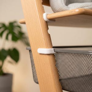 May include: Close-up of a wooden high chair with a light brown finish. The chair has a shelf and a metal basket. White plastic clips secure the shelf and basket to the wooden frame. A grey cushion is visible on the top shelf.