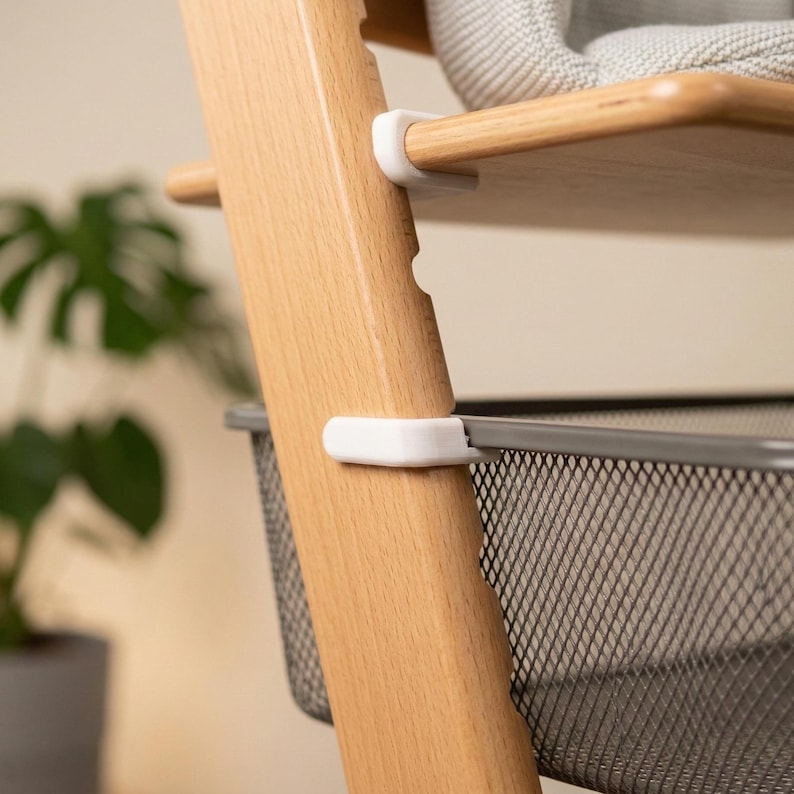 May include: Close-up of a wooden high chair with a light brown finish. The chair has a shelf and a metal basket. White plastic clips secure the shelf and basket to the wooden frame. A grey cushion is visible on the top shelf.