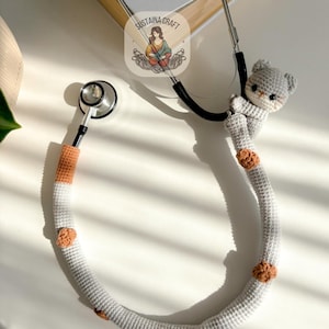 May include: A gray crochet stethoscope cover with a small gray bear attached. The cover has orange and brown accents and is designed to fit a standard stethoscope. The stethoscope rests on a white surface.