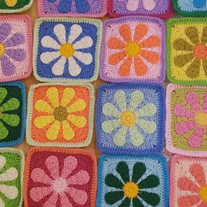 May include: A vibrant, handmade crochet blanket with a repeating pattern of square blocks. Each block features a flower design in various colours, including pink, yellow, blue, and green. The blanket is a patchwork of floral motifs.