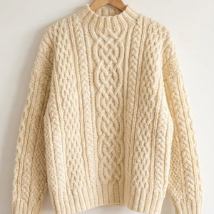 May include: Cream-colored cable-knit sweater with a high neckline. The sweater features intricate cable patterns down the front and sleeves, with ribbed detailing at the cuffs and hem. The garment is displayed on a wooden hanger.