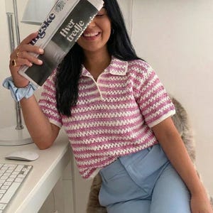 Op de afbeelding: Een roze en wit gestreept gehaakt poloshirt met een witte kraag en korte mouwen. Het shirt wordt gecombineerd met een lichtblauwe broek. De afbeelding bevat ook een krant met de tekst "Hver tredje".