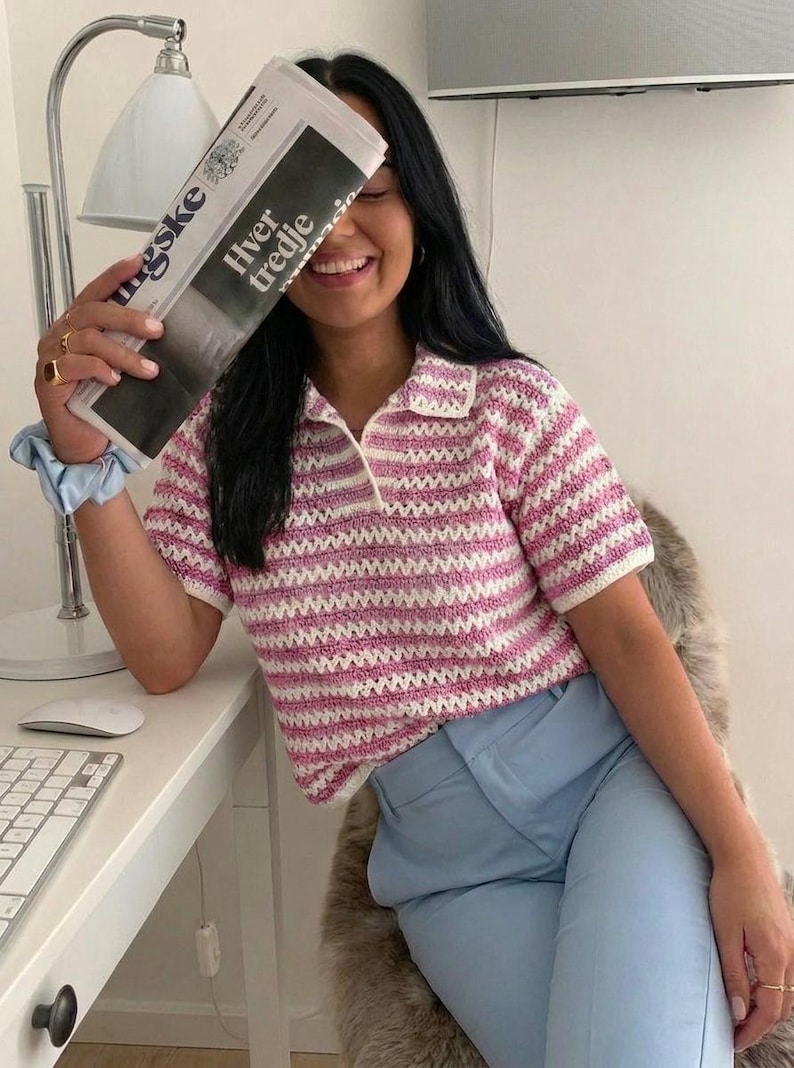 Op de afbeelding: Een roze en wit gestreept gehaakt poloshirt met een witte kraag en korte mouwen. Het shirt wordt gecombineerd met een lichtblauwe broek. De afbeelding bevat ook een krant met de tekst "Hver tredje".