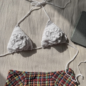 Puede incluir: Top de bikini blanco de crochet con detalles florales. La parte superior se ata en el cuello y la espalda. La parte inferior es de tela estampada de colores.