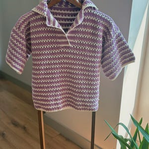 Op de afbeelding: Een gehaakt shirt met korte mouwen in een lavendel- en wit gestreept patroon. Het shirt heeft een kraag en wordt gepresenteerd op een houten hanger. Het open geweven ontwerp is zichtbaar in het hele kledingstuk, waardoor het een lichte en luchtige uitstraling heeft.