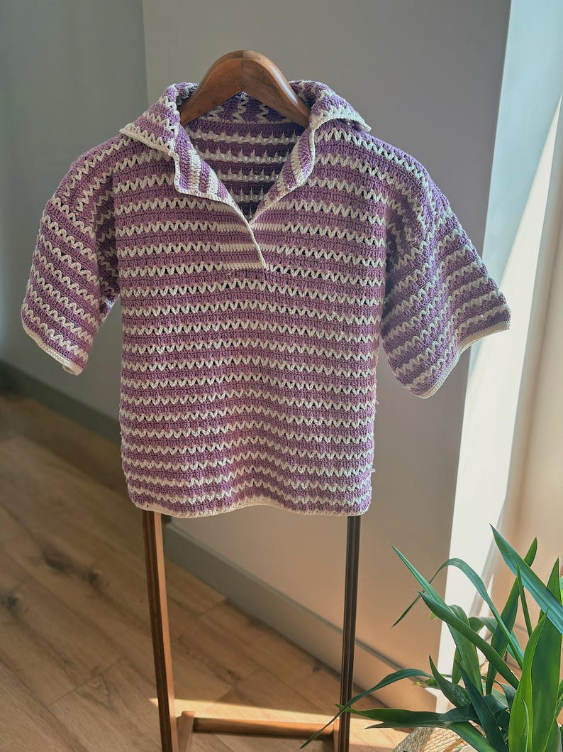 Op de afbeelding: Een gehaakt shirt met korte mouwen in een lavendel- en wit gestreept patroon. Het shirt heeft een kraag en wordt gepresenteerd op een houten hanger. Het open geweven ontwerp is zichtbaar in het hele kledingstuk, waardoor het een lichte en luchtige uitstraling heeft.