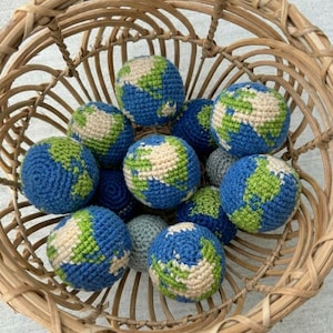 May include: A collection of crocheted globe-shaped balls in a woven basket. The balls feature blue oceans, green continents, and beige landmasses. The basket is made of light brown woven material.