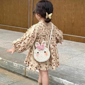Pode incluir: Uma criança vestindo um vestido com estampa floral e uma bolsa de coelho de crochê branco. O vestido tem mangas compridas e gola alta. A bolsa tem um laço rosa e olhos pretos. A criança também está usando sapatos e meias marrons.