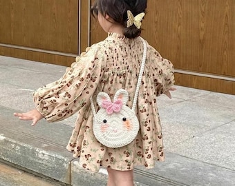 Könnte beinhalten: Ein Kind trägt ein geblümtes Kleid mit einer weißen Häkeltasche in Hasenform. Das Kleid hat lange Ärmel und einen hohen Ausschnitt. Die Tasche hat eine rosa Schleife und schwarze Augen. Das Kind trägt auch braune Schuhe und Socken.