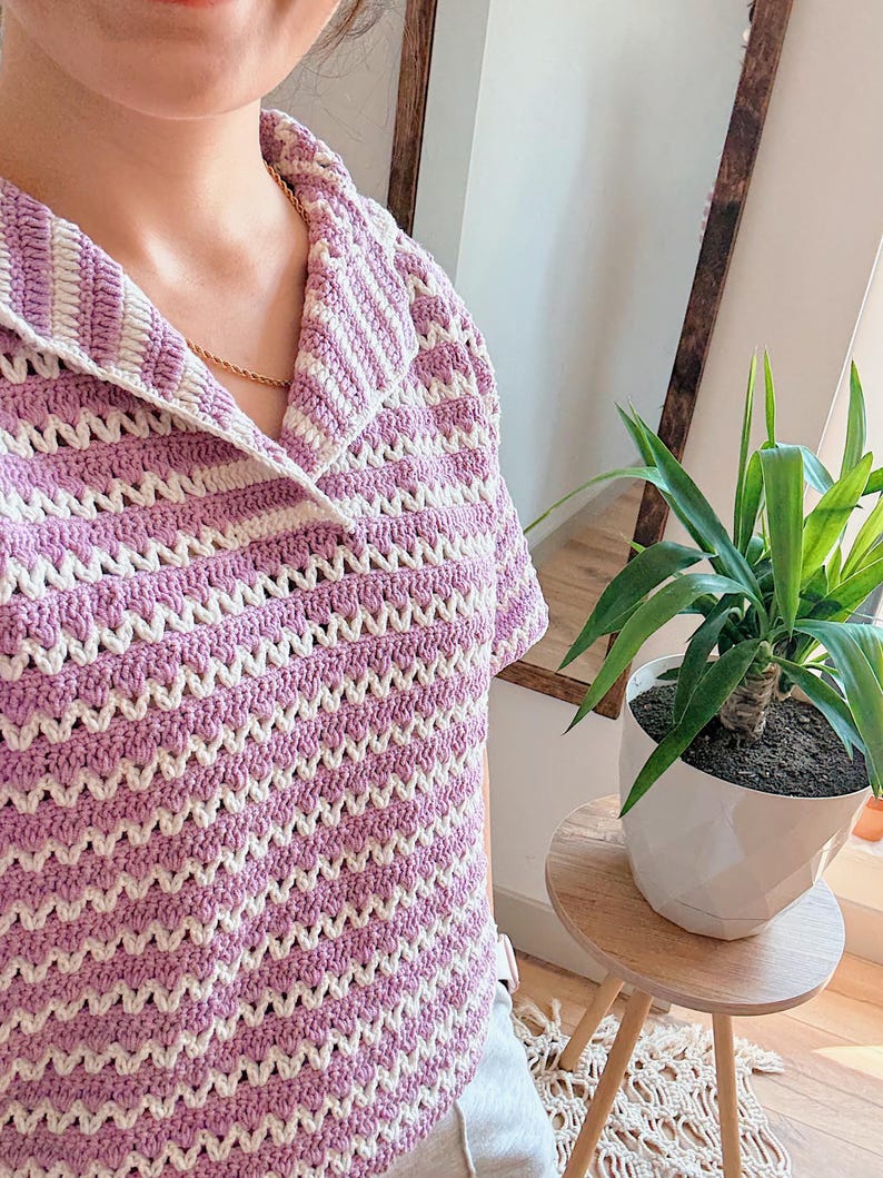 Op de afbeelding: Een gehaakte top met korte mouwen in een lavendel- en wit chevronpatroon. De top heeft een kraag en een relaxte pasvorm. Het handgemaakte kledingstuk is een stijlvolle toevoeging aan elke garderobe.