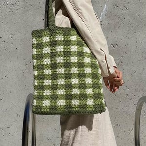 Könnte beinhalten: Eine grüne und weiße Gingham-Tragetasche mit einem gehäkelten Design. Die Tasche wird von einer Person getragen, die ein beiges Hemd trägt.