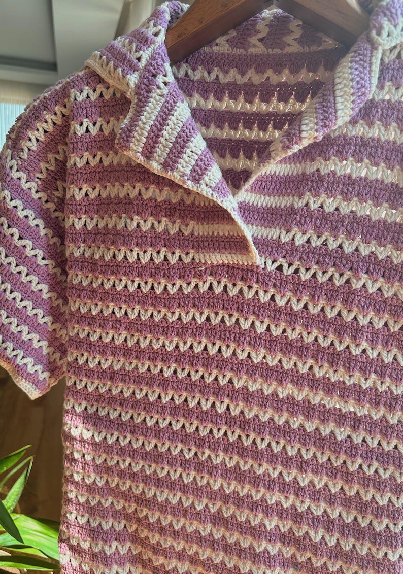 Op de afbeelding: Een gehaakt shirt met korte mouwen in een lavendel- en wit gestreept patroon. Het shirt heeft een kraag en een knoopsluiting. Het haakontwerp cre&euml;ert een open, luchtige textuur, perfect voor warm weer.