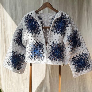 Può includere: Un cardigan bianco lavorato a uncinetto con dettagli blu e rosa. Il cardigan ha un motivo quadrato e una vestibilità ampia.