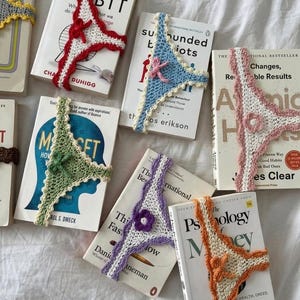 May include: A collection of seven crochet bookmarks shaped like thong underwear, each featuring a different color trim including red, blue, pink, green, purple, orange, and yellow, placed over various book covers with visible titles and text.