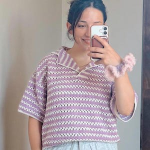 Op de afbeelding: Een gehaakte top met korte mouwen in een zigzagpatroon van lavendel en wit. De top heeft een kraag en een relaxte pasvorm. De mouwen en kraag zijn wit afgewerkt. Een roze en lavendelkleurige scrunchie is zichtbaar.