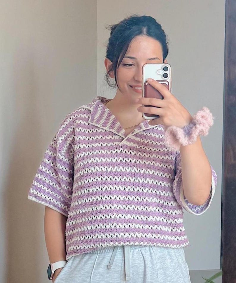 Op de afbeelding: Een gehaakte top met korte mouwen in een zigzagpatroon van lavendel en wit. De top heeft een kraag en een relaxte pasvorm. De mouwen en kraag zijn wit afgewerkt. Een roze en lavendelkleurige scrunchie is zichtbaar.