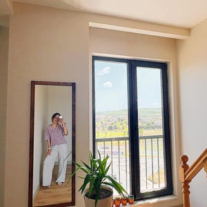 Op de afbeelding: Een lange spiegel met een donkerbruin houten frame hangt aan een beige muur. De spiegel reflecteert een persoon die een gestreept shirt en een lichte broek draagt. Rechts is een groot raam met uitzicht op een groen landschap.