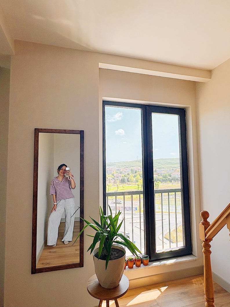 Op de afbeelding: Een lange spiegel met een donkerbruin houten frame hangt aan een beige muur. De spiegel reflecteert een persoon die een gestreept shirt en een lichte broek draagt. Rechts is een groot raam met uitzicht op een groen landschap.