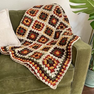 Może przedstawiać: Ręcznie robiony koc z dzianiny w stylu granny square w odcieniach brązu, zieleni, pomarańczy i bieli. Koc leży na zielonej kanapie.