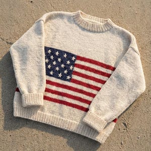 Op de afbeelding: Crèmekleurige gebreide trui met een Amerikaans vlagontwerp. De trui heeft een ronde hals, lange mouwen en geribde manchetten en zoom. Het vlagontwerp bevat een blauw veld met witte sterren en rode en witte strepen.