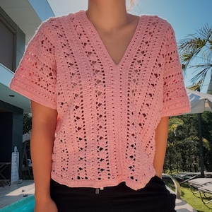 Peut inclure: Un haut en crochet rose clair avec un col en V et des manches courtes. Le motif ajouré crée un aspect délicat et aéré. Le haut est fait d'un fil doux et texturé, parfait pour les journées chaudes. Le haut est de coupe ample.