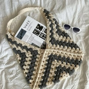 May include: A crochet hobo bag featuring a geometric granny square pattern in alternating cream and charcoal gray yarn. The bag contains a magazine with the text VOGUE IN COPENHAGEN visible on the cover, accompanied by white oval sunglasses.
