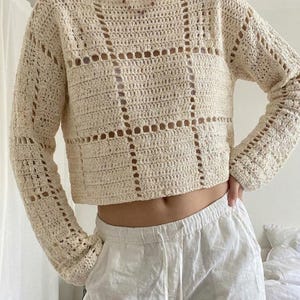 May include: Cream-colored, long-sleeved crochet top with a square pattern. The top has a wide neckline and is paired with white drawstring pants. The crochet design features open squares and a cropped length.