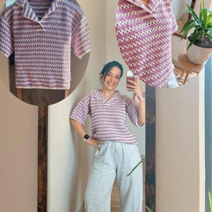 Op de afbeelding: Een gebreide top met korte mouwen en kraag in een lavendel- en wit gestreept patroon. De top wordt op een hanger en door een persoon gedragen. De persoon draagt ​​grijze joggingbroek. De top heeft een relaxte pasvorm.