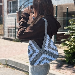 Peut inclure: Un sac cabas en crochet gris et blanc avec un motif carré. Le sac est porté sur l'épaule d'une personne vêtue d'un pull marron et d'un pantalon gris.