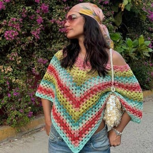 Könnte beinhalten: Ein mehrfarbiger Häkelponcho mit V-förmigem Muster und Streifen aus rotem, gelbem, grünem und blauem Garn. Das Kleidungsstück hat eine grobmaschige Textur und einen weiten Ausschnitt. Eine weiße Pailletten-Umhängetasche wird über dem Poncho getragen.
