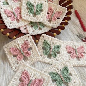 May include: Handmade crochet butterfly squares in white, pink, and green. The squares are arranged in a wooden basket. The butterflies are centered on the squares. The squares are a decorative craft item.