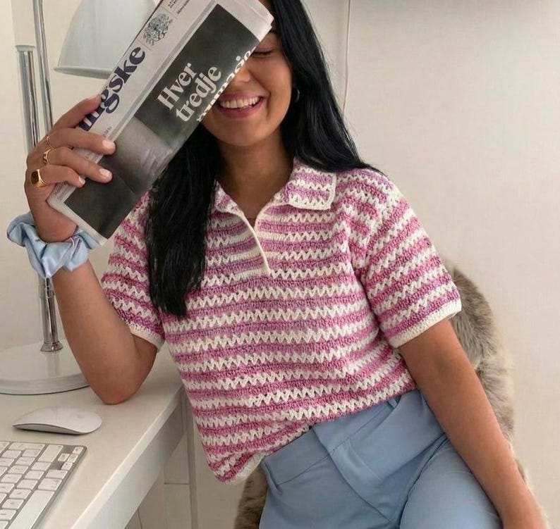 Op de afbeelding: Een roze en wit gestreepte gehaakte poloshirt met een witte kraag en korte mouwen. Het shirt wordt gecombineerd met lichtblauwe broek. Een krant wordt dicht bij het gezicht gehouden.
