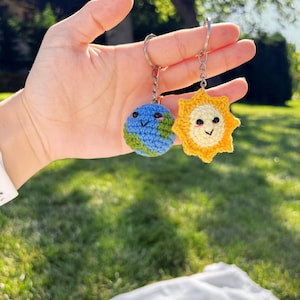 Pode incluir: Dois chaveiros de crochê: um representando a Terra com detalhes azuis e verdes, e o outro um sol amarelo com um rosto sorridente. Ambos os chaveiros são presos a argolas prateadas, perfeitos para adicionar um toque de fantasia.