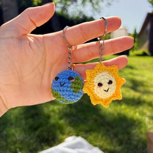 May include: Two crochet keychains: one blue and green Earth and one yellow sun, both with smiling faces. The keychains are attached to silver key rings. The background is a blurred outdoor scene with green grass and trees.