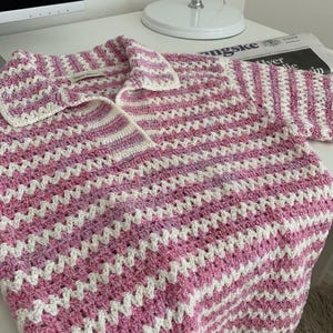 Op de afbeelding: Een handgemaakt gehaakt shirt in een roze en wit chevronpatroon. Het shirt heeft een kraag en lange mouwen. Het haakwerk is gedetailleerd en de kleuren zijn zacht. Het shirt ligt op een wit oppervlak.