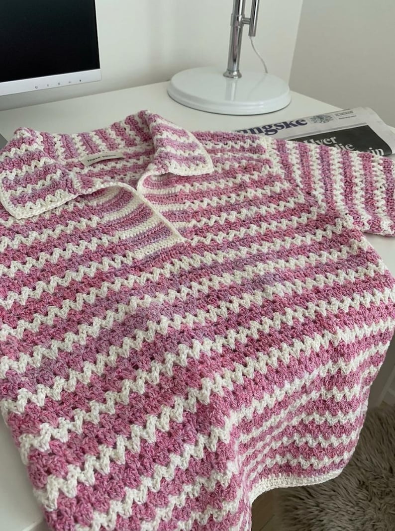 Op de afbeelding: Een handgemaakt gehaakt shirt in een roze en wit chevronpatroon. Het shirt heeft een kraag en lange mouwen. Het haakwerk is gedetailleerd en de kleuren zijn zacht. Het shirt ligt op een wit oppervlak.