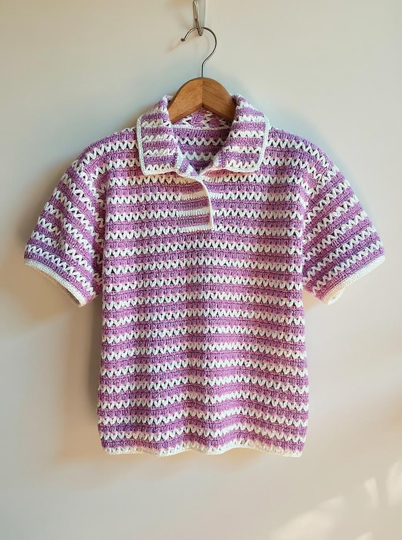 Op de afbeelding: Een gehaakte poloshirt met korte mouwen in een gestreept patroon van lavendel en wit. De shirt heeft een kraag, een knoopsluiting met drie knopen en witte afwerking op de kraag en mouwen. De shirt hangt aan een houten hanger.