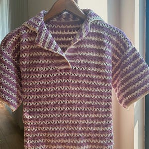 Op de afbeelding: Een gehaakt shirt met korte mouwen in een lavendel- en wit gestreept patroon. Het shirt heeft een kraag en wordt gepresenteerd op een houten hanger. Het open weefselontwerp is zichtbaar in het hele kledingstuk.