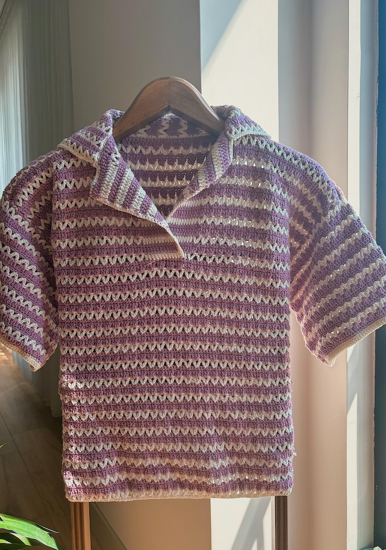 Op de afbeelding: Een gehaakt shirt met korte mouwen in een lavendel- en wit gestreept patroon. Het shirt heeft een kraag en wordt gepresenteerd op een houten hanger. Het open weefselontwerp is zichtbaar in het hele kledingstuk.