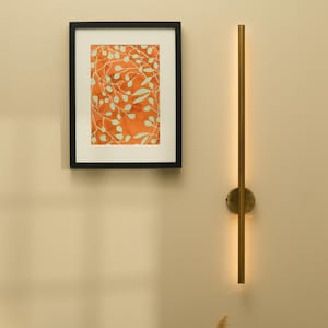 May include: A framed artwork with an orange background and a white leaf pattern hangs on a beige wall. A gold-colored, vertical wall sconce with a warm light is mounted next to the artwork. A black side table holds decorative items.