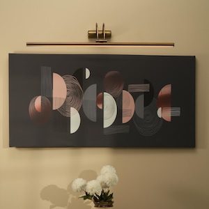 May include: A modern abstract art piece with geometric shapes in shades of gray, white, and rose gold hangs above a small black table. A cream-colored vase with white flowers sits on the table, illuminated by a brass-toned picture light.