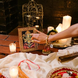 Puede incluir: Una impresi&oacute;n enmarcada de la deidad Ganesha en un marco dorado, acompa&ntilde;ada de una tela blanca, un plato decorativo rojo con cuencos de metal, una jaula de p&aacute;jaros de metal, varias velas blancas encendidas y una guirnalda de cuentas naranjas.
