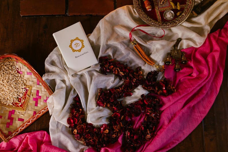 May include: A white book titled Bhagavad Gita with an orange Om symbol, a woven tray with grains, a pink and white fabric, a dark red petal arrangement in an Om shape, a beaded string, and a small metallic bell ornament on a dark surface.